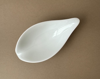 Könnte beinhalten: Weiße Keramik-Servierplatte in Blattform. Die Platte ist flach und hat eine glatte, glänzende Oberfläche.