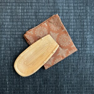 May include: A light brown, oval-shaped, wooden dish with a slightly raised edge. The dish is sitting on a brown and white patterned fabric.