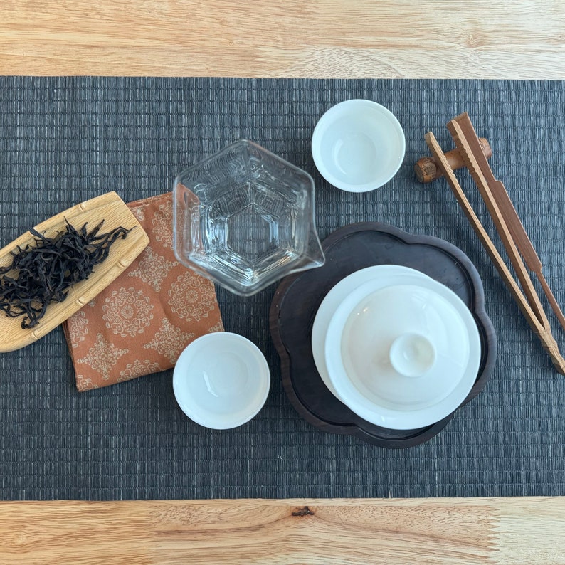 May include: A black tea leaf set with a glass teapot, a white teacup, a wooden tea tong, and a brown and white patterned tea towel.