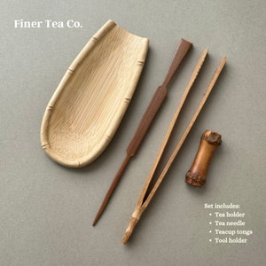 May include: A set of four wooden tea tools, including a tea holder, tea needle, teacup tongs, and a tool holder. The tools are made of natural bamboo and wood, and are arranged on a light gray background. The text "Finer Tea Co." is visible at the top of the image.