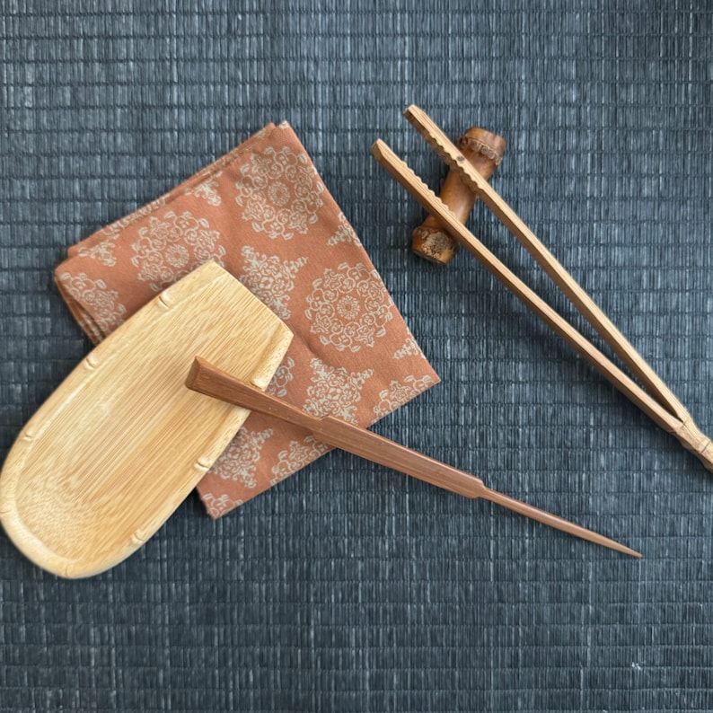 May include: A set of wooden kitchen utensils, including a small serving dish, a pair of chopsticks, and a set of tongs. The utensils are arranged on a brown fabric background.