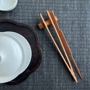 May include: Two wooden tea tongs with a natural finish. The tongs are on a dark gray woven mat.