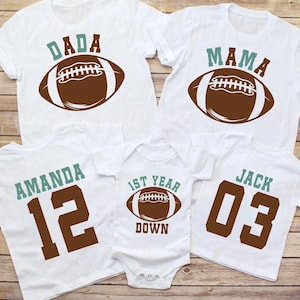 May include: White t-shirts with football-themed designs. The shirts feature the words "DADA," "MAMA," "AMANDA 12," and "JACK 03," along with a football graphic. A onesie with "1st YEAR DOWN" and a football graphic is also included.