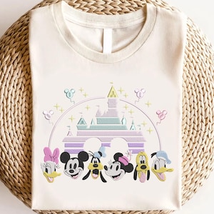 May include: A white t-shirt with an embroidered design featuring Mickey Mouse, Minnie Mouse, Donald Duck, Goofy, and Pluto in front of a pastel purple and green version of the Cinderella Castle.  The design also includes pastel balloons and stars.