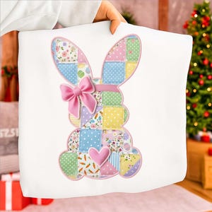 Könnte beinhalten: Weißes Kissen mit einem Patchwork-Hasen-Design. Der Hase hat eine rosa Schleife, ein rosa Herz und Ohren mit Blumen- und Tupfenmustern. Das Design ist rosa umrandet.