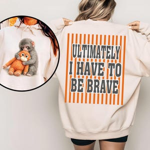 Op de afbeelding: Crèmekleurige sweatshirt met een afbeelding van een aap die een knuffelbeest omhelst op de voorkant. De achterkant van de sweatshirt toont de tekst "ULTIMATELY I HAVE TO BE BRAVE" in een vette letter, omlijst door verticale oranje strepen.