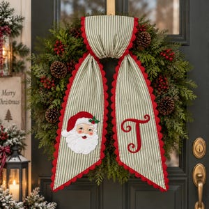 May include: A green evergreen wreath decorated with pinecones and red berries, featuring a hanging fabric banner with green and white vertical stripes, red scalloped edges, an embroidered Santa Claus face, and a large red embroidered letter T.