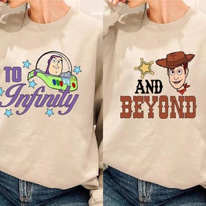Puede incluir: Dos sudaderas beige con diseños gráficos. Una presenta a Buzz Lightyear en una nave espacial con el texto "To Infinity" en morado. La otra muestra a Woody con una placa de sheriff y el texto "And Beyond" en marrón.