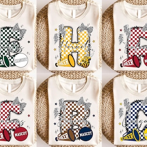 May include: Six cream-colored t-shirts with different team names and mascots printed on them. Each shirt features a large letter, a megaphone, and the word "CHEER". The designs include checkered patterns, stars, and lightning bolts. The team names are Eagles, Hornets, Falcons, Bobcats, and Cougars.
