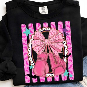 May include: A black long-sleeve sweatshirt featuring a graphic print of two pink boxing gloves tied together with a large pink bow patterned with breast cancer awareness ribbons, set against a background of pink bat-patterned stripes and stars.