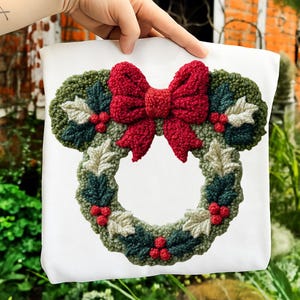 May include: White tote bag featuring a Christmas wreath design. The wreath is shaped like Mickey Mouse ears and includes a red bow, green leaves, and red berries. The design is made of textured fabric.