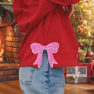 May include: A red sweatshirt with a pink glitter bow on the back. The sweatshirt is worn over blue jeans.