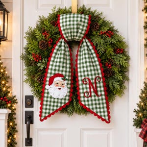 Peut inclure: Une couronne de Noël composée de branches de cèdre vertes, de pommes de pin et de baies rouges. Un ruban en tissu vichy vert et blanc à bords festonnés rouges pend du haut, orné d'un visage de Père Noël brodé et de la lettre N.