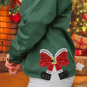 May include: A green sweatshirt with a sparkly red and white Santa bow on the back. The bow is made of glitter and has a black belt buckle.