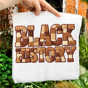 May include: White sweatshirt with the words "BLACK HISTORY" in large, textured, brown and tan letters. The letters have a checkerboard and striped pattern. The sweatshirt is held up by a hand.