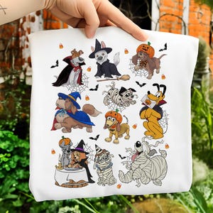 May include: A white tote bag featuring a colorful graphic print of various cartoon dogs dressed in Halloween costumes, including vampires, mummies, and witches, surrounded by small bats, spider webs, and candy corn illustrations.
