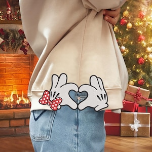 May include: Beige hoodie with a heart-shaped hand gesture design. The design features white gloves with black outlines and a red polka dot bow. The hoodie is paired with blue jeans, creating a casual and festive look.