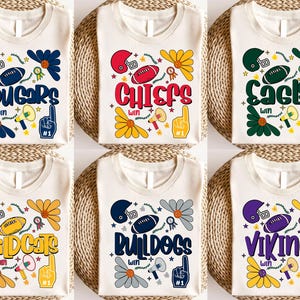 Pode incluir: Seis camisetas creme com nomes de equipes e gráficos. Cada camisa apresenta um capacete de futebol, uma bola de futebol, uma flor, um megafone e uma mão com o número um. Os nomes das equipes incluem Cougars, Chiefs, Eagles, Wildcats, Bulldogs e Vikings.