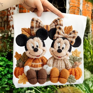 May include: A white pillow featuring embroidered Mickey and Minnie Mouse characters in fall attire. Mickey wears an orange sweater, while Minnie has a beige sweater and bow. Pumpkins and fall leaves surround the characters, creating a seasonal theme.