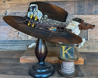 May include: A brown cowboy hat with a wide brim, decorated with barbed wire, a skull, and a photo. The hat is displayed on a dark wooden stand. A wooden block with the letter "K" is next to the stand.