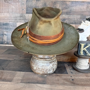 Peut inclure: Chapeau fedora vert olive vieilli avec un bandeau en cuir marron et des coutures orange. Le chapeau a un large bord et est orné d'une plume et d'une petite douille en laiton. Le chapeau est présenté sur un présentoir en bois décoratif.