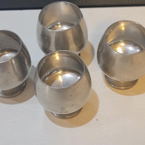 May include: Four silver shot glasses with a rounded bottom and a wide rim. The glasses are arranged in a semi-circle on a white surface.