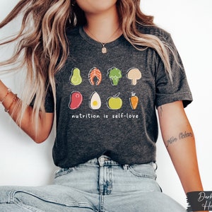 Puede incluir: Camiseta de color gris oscuro jaspeado con un gr&aacute;fico de varios alimentos saludables y el texto "nutrition is self-love".
