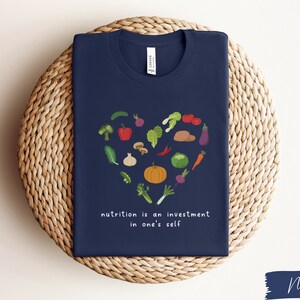 Pu&ograve; includere: Maglietta blu navy con un design a forma di cuore fatto di varie verdure e il testo "nutrition is an investment in one's self".