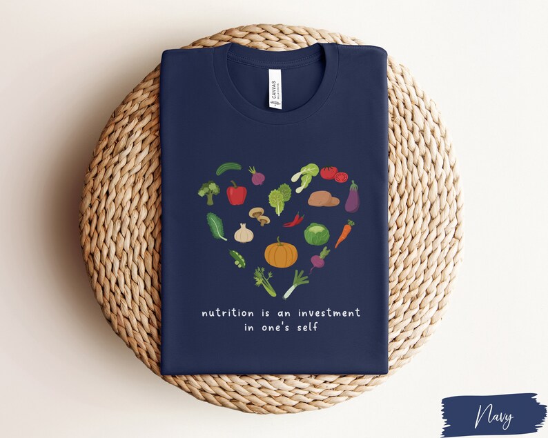 Pu&ograve; includere: Maglietta blu navy con un design a forma di cuore fatto di varie verdure e il testo "nutrition is an investment in one's self".