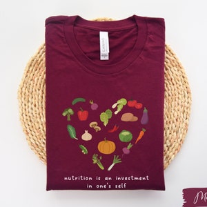 Pu&ograve; includere: Maglietta color marsala con un design a forma di cuore fatto di verdure colorate e il testo "nutrition is an investment in one's self".