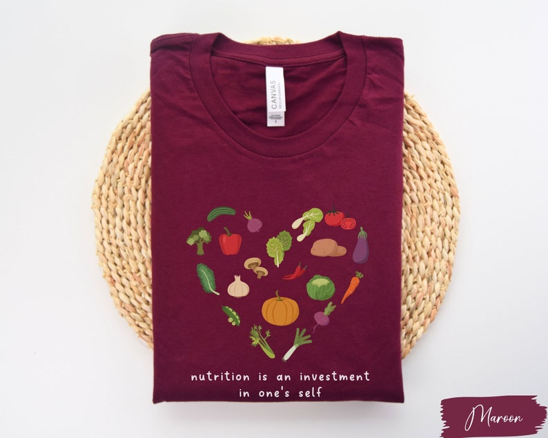 Pu&ograve; includere: Maglietta color marsala con un design a forma di cuore fatto di verdure colorate e il testo "nutrition is an investment in one's self".