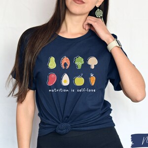 Puede incluir: Una camiseta azul marino con un gr&aacute;fico de varias frutas y verduras. El texto "nutrition is self-love" est&aacute; impreso debajo del gr&aacute;fico.