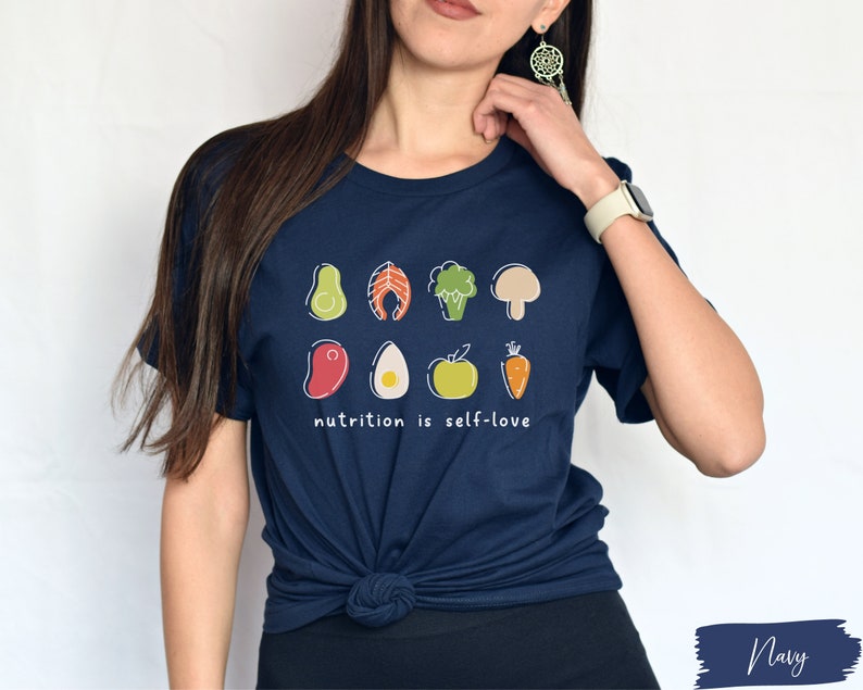Puede incluir: Una camiseta azul marino con un gr&aacute;fico de varias frutas y verduras. El texto "nutrition is self-love" est&aacute; impreso debajo del gr&aacute;fico.