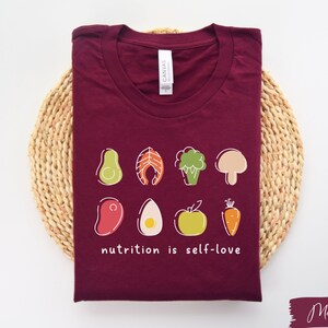 Puede incluir: Camiseta de color granate con un gr&aacute;fico blanco de varios alimentos y el texto "nutrition is self-love".