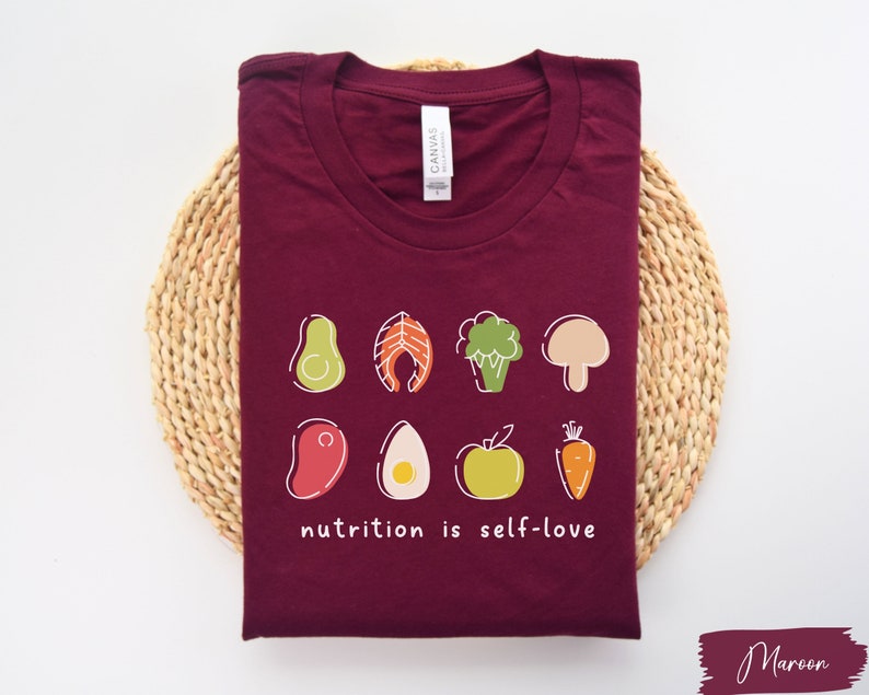 Puede incluir: Camiseta de color granate con un gr&aacute;fico blanco de varios alimentos y el texto "nutrition is self-love".