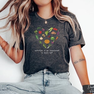 Pu&ograve; includere: Maglietta in jersey grigio scuro con un design a forma di cuore fatto di varie verdure e il testo "nutrition is an investment in one's self".