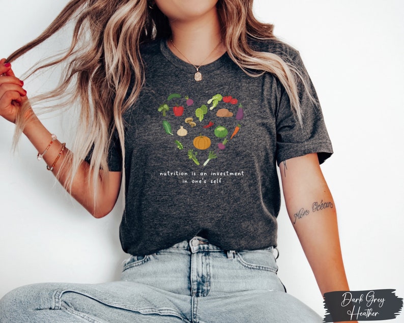 Pu&ograve; includere: Maglietta in jersey grigio scuro con un design a forma di cuore fatto di varie verdure e il testo "nutrition is an investment in one's self".