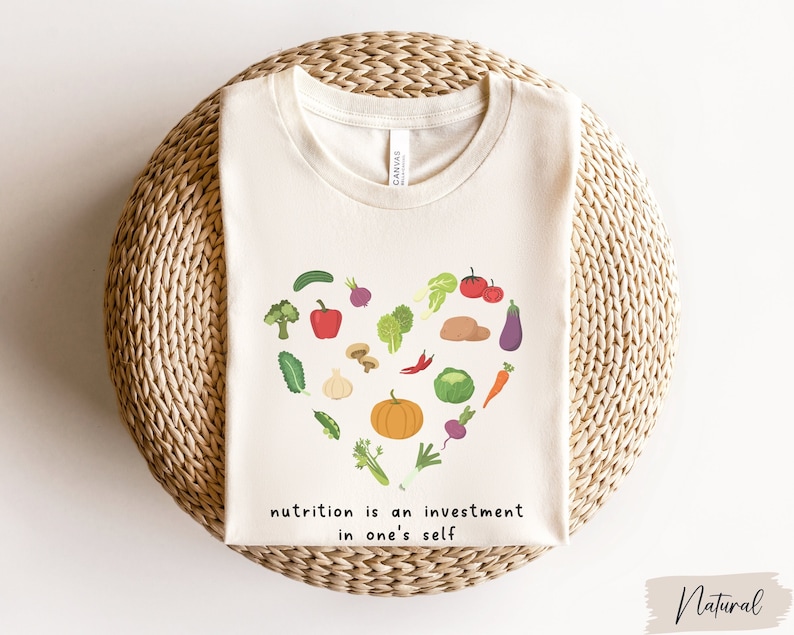 Pu&ograve; includere: Una maglietta bianca con un design a forma di cuore fatto di varie verdure e frutta. Il testo "nutrition is an investment in one's self" &egrave; stampato sotto il design.