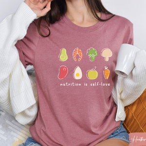 Puede incluir: Una camiseta de color malva con un gr&aacute;fico de varios alimentos saludables, incluyendo un aguacate, pescado, br&oacute;coli, champi&ntilde;&oacute;n, huevo, manzana y zanahoria. El texto "nutrition is self-love" est&aacute; impreso debajo del gr&aacute;fico.