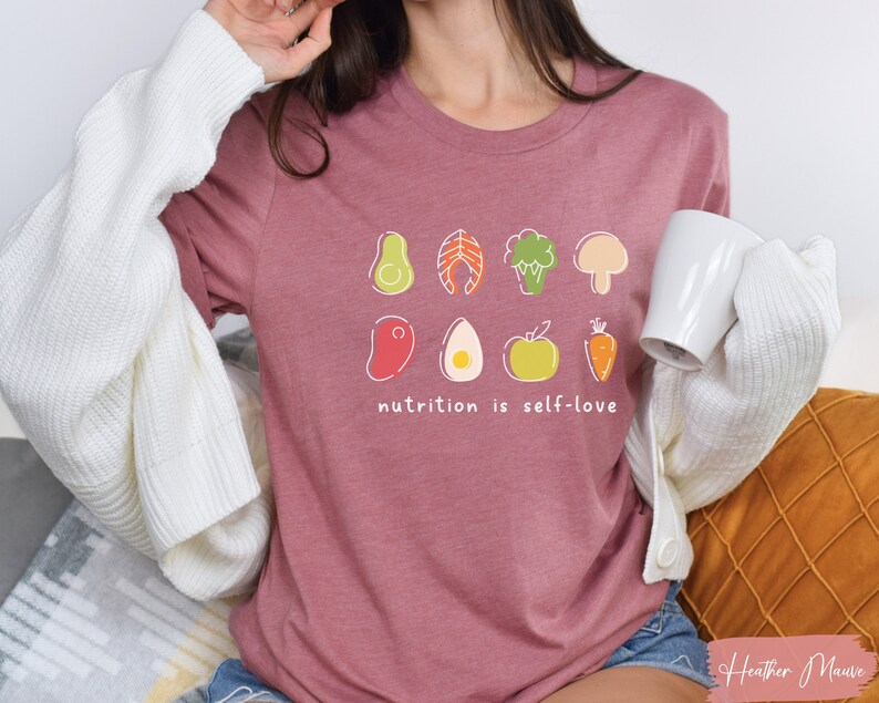 Puede incluir: Una camiseta de color malva con un gr&aacute;fico de varios alimentos saludables, incluyendo un aguacate, pescado, br&oacute;coli, champi&ntilde;&oacute;n, huevo, manzana y zanahoria. El texto "nutrition is self-love" est&aacute; impreso debajo del gr&aacute;fico.