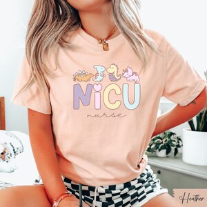 Puede incluir: Una camiseta rosa con un gr&aacute;fico de dinosaurios coloridos y el texto "NICU nurse".