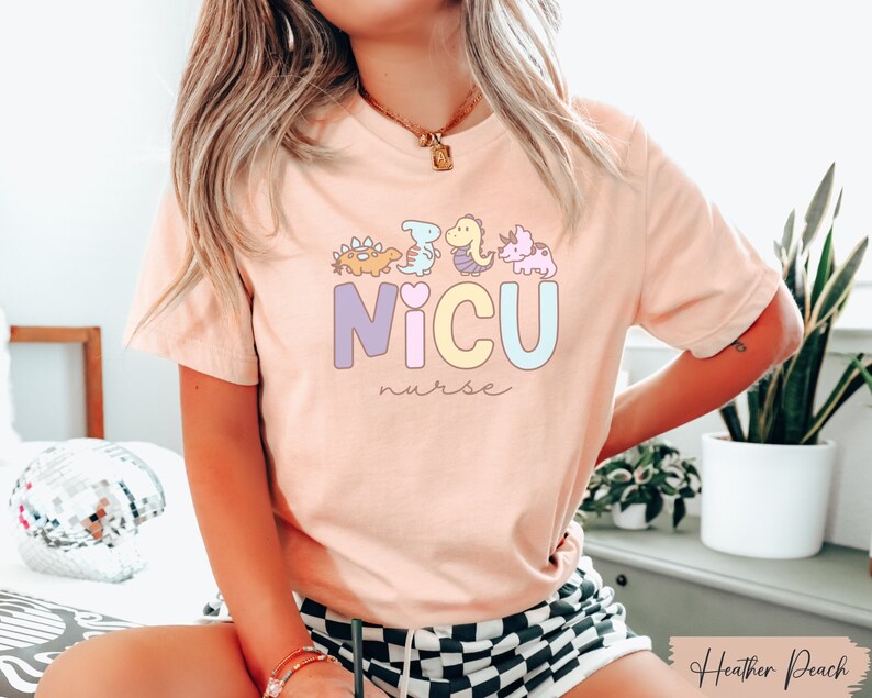 Puede incluir: Una camiseta rosa con un gr&aacute;fico de dinosaurios coloridos y el texto "NICU nurse".