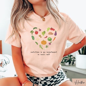 Pu&ograve; includere: Una maglietta rosa con un design a forma di cuore fatto di varie verdure e il testo "nutrition is an investment in one's self".