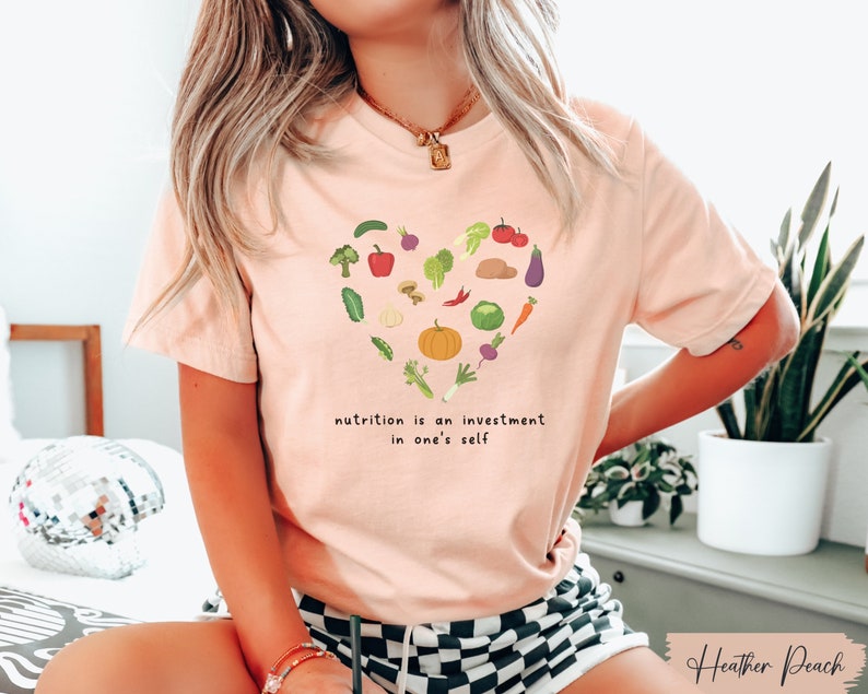 Pu&ograve; includere: Una maglietta rosa con un design a forma di cuore fatto di varie verdure e il testo "nutrition is an investment in one's self".