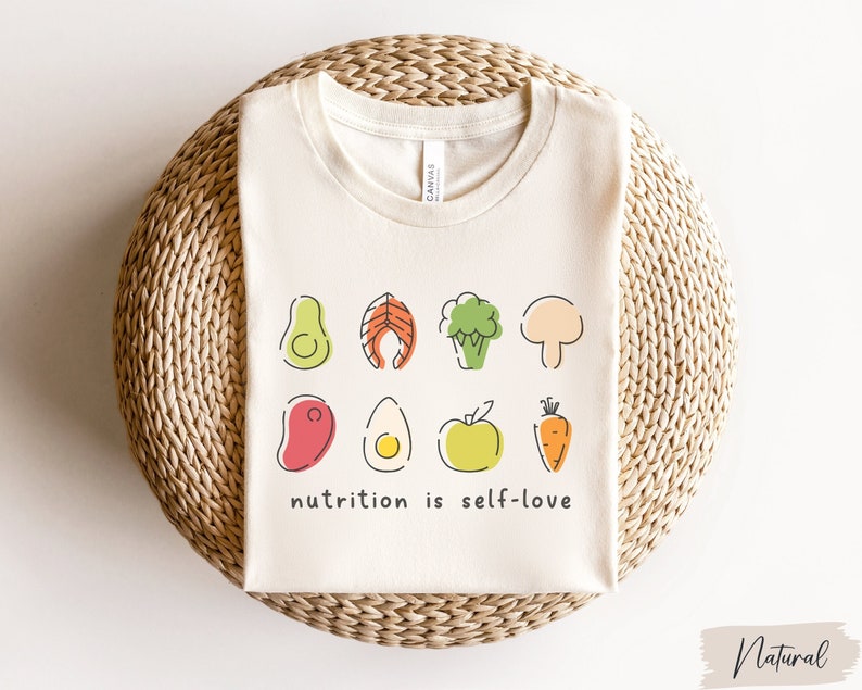 Puede incluir: Una camiseta beige con un gr&aacute;fico de varios alimentos, incluyendo un aguacate, salm&oacute;n, br&oacute;coli, champi&ntilde;&oacute;n, bistec, huevo, manzana y zanahoria. El texto "nutrition is self-love" est&aacute; impreso debajo del gr&aacute;fico.