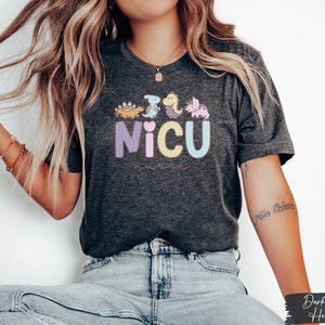 Puede incluir: Una camiseta de color gris oscuro jaspeado con un gr&aacute;fico de dinosaurios coloridos y el texto "NICU Nurse".