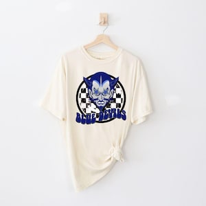 May include: A cream colored t-shirt with a blue and white graphic of a devil's face with the text "Blue Devils" on a checkered background.