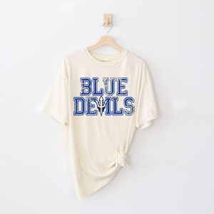 May include: Cream-colored t-shirt with the text "BLUE DEVILS" in blue block letters. The shirt is hanging on a wooden hanger against a white background. The text has a distressed, vintage look.