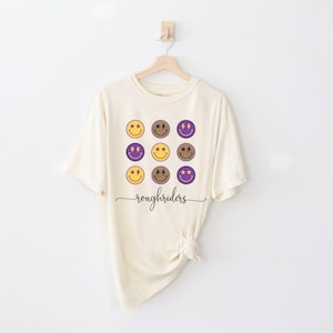 May include: A cream-colored t-shirt with a graphic of nine purple and gold smiley faces and the text "roughriders".