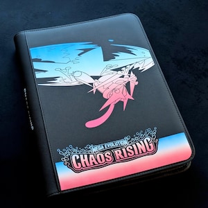 May include: Black binder with a zip closure, featuring a graphic of a creature and the text "MEGA EVOLUTION CHAOS RISING". The design incorporates pink, blue, and white colours. Likely for storing trading cards or collectibles.